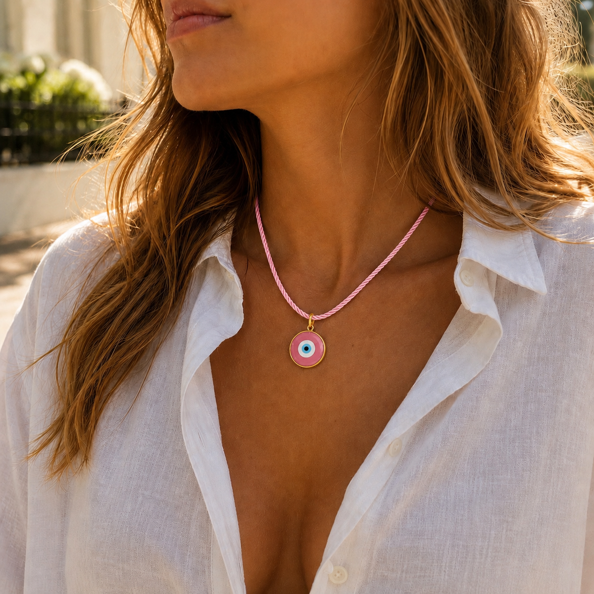 A woman wearing a pink beaded necklace with a circular evil eye pendant, styled with an open white linen shirt in a sunny outdoor setting.